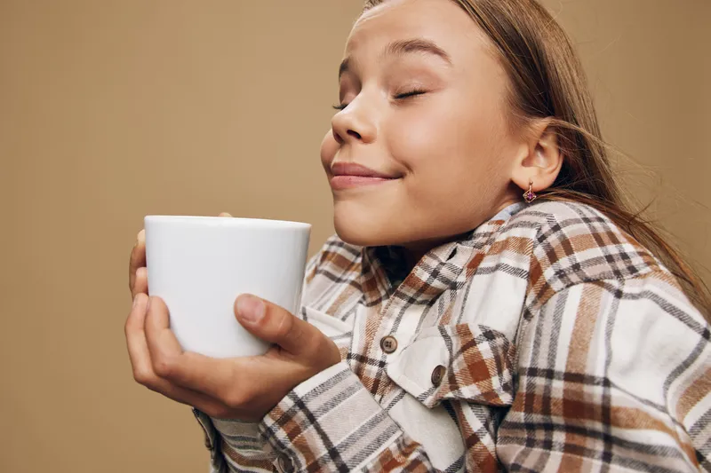 Young woman holding white cup with closed eyes and smile, enjoying aroma and warmth. Casual clothing, cozy atmosphere and light beige background with relaxed feeling.