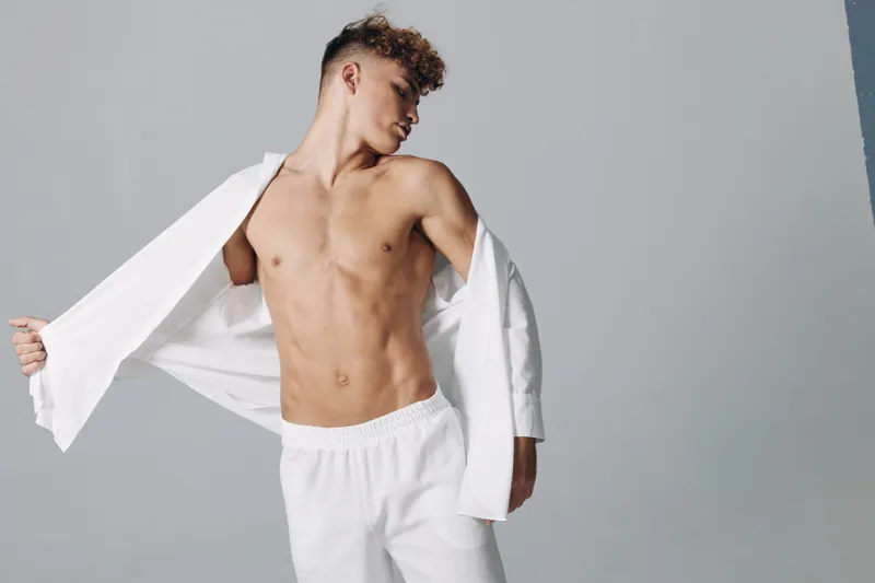 Young man with muscular body posing shirtless while putting on white shirt against plain background. Fitness, fashion and casual style with confident expression and modern look.