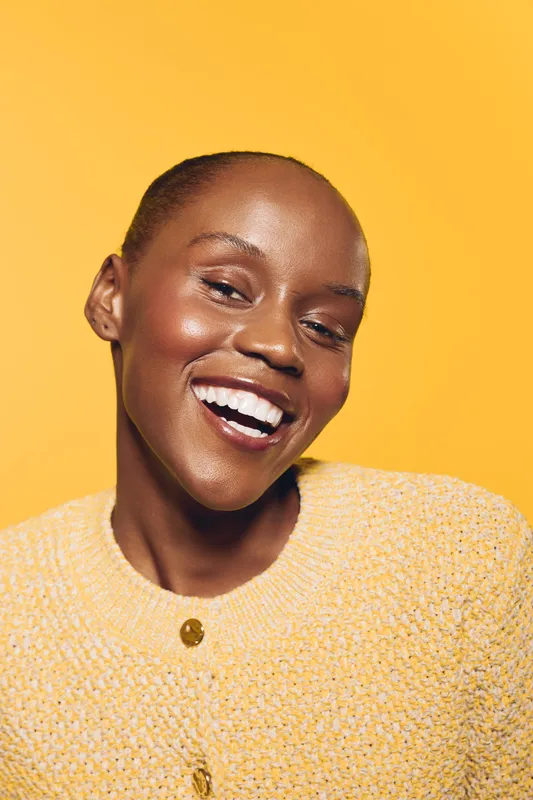 Smiling African American woman with dark skin and natural makeup wearing a yellow sweater. Close-up portrait on yellow background. Concept of happiness, beauty, confidence, and wellness.