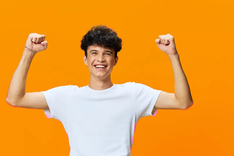 Happy man with raised fists celebrating success wearing a white t-shirt on bright orange background expressing excitement and energy.