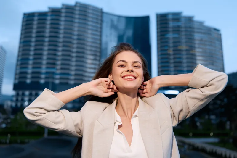 Confident smiling woman in beige blazer enjoying city skyscraper background, bright natural light, urban business fashion concept