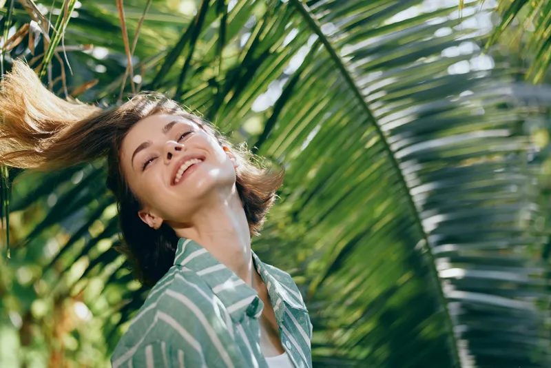 Bright, carefree portrait of a woman smiling under tropical palm fronds, sunlight streaming through leaves, conveying joy and natural beauty.