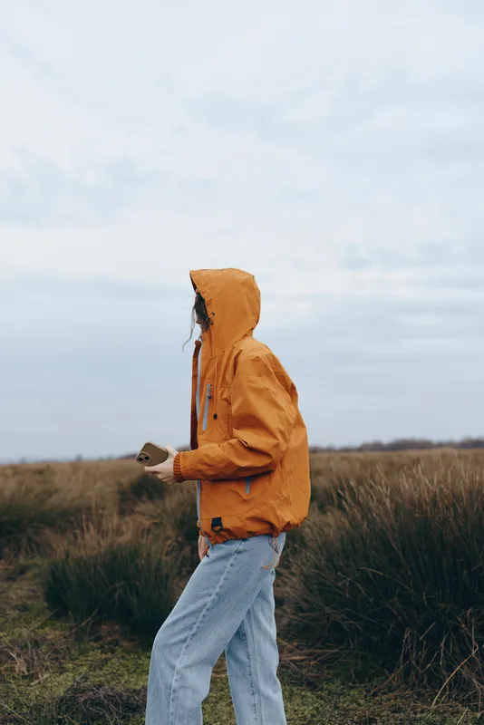 Woman outdoors lifestyle, hooded jacket in a grassy field, walking in wind under cloudy sky, relaxed casual posture, warm orange color, soft natural light, wide horizon, quiet countryside
