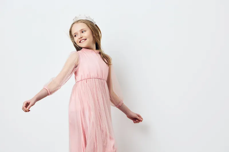 Young girl in a pink dress with a crown, joyfully posing against a light background, evoking elegance and innocence in a springtime setting