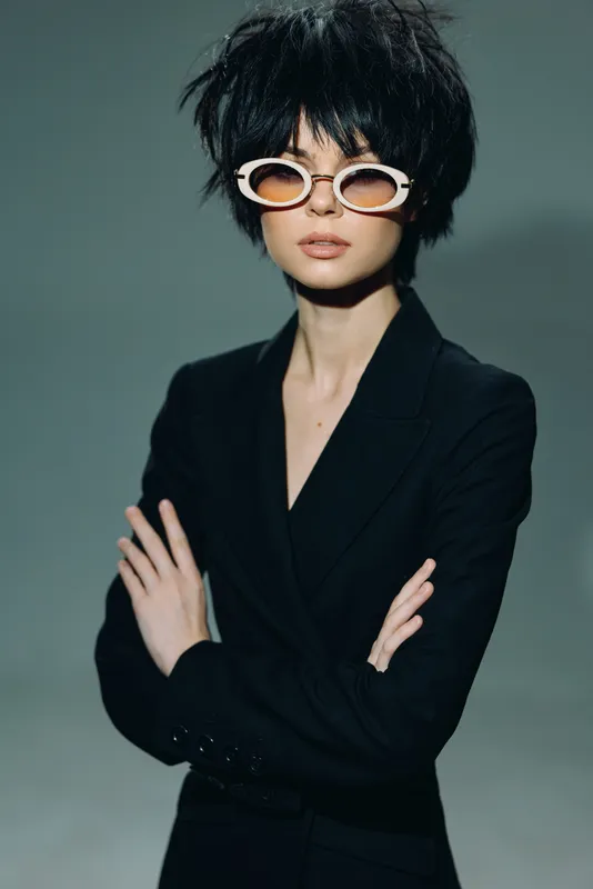 Stylish woman with short black hair wearing sunglasses and a black blazer against a gray background.