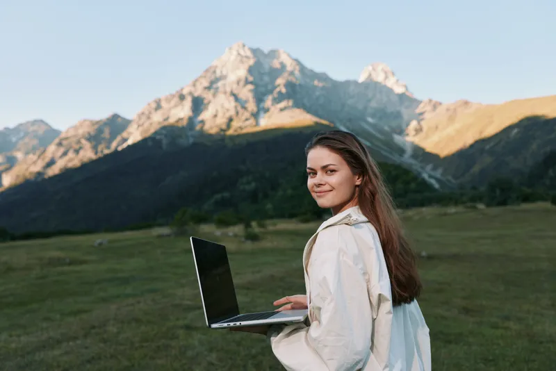 A cheerful woman works outdoors with a laptop in a scenic meadow beneath mountains. She smiles at the screen, projecting focus, enthusiasm, and a vibrant, modern work vibe.