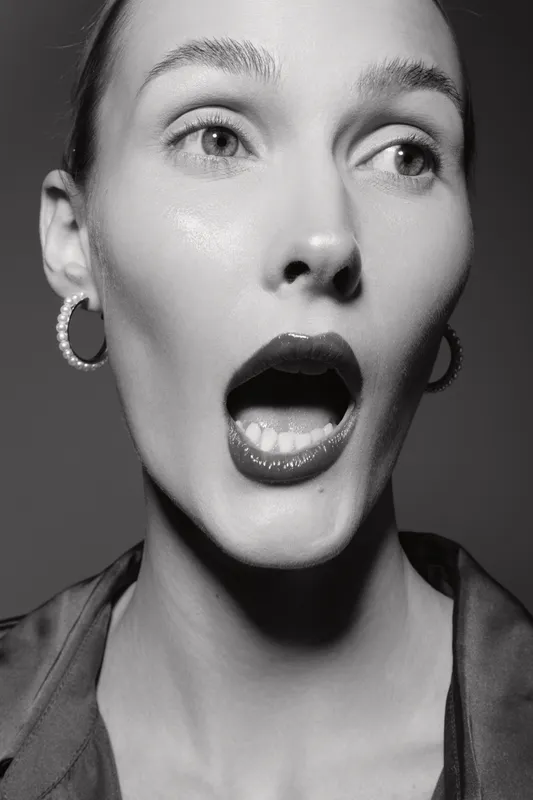 Close-up of a surprised Caucasian woman with light skin and wide eyes, mouth open in shock. Studio portrait in black and white with hoop earrings and slicked hair.