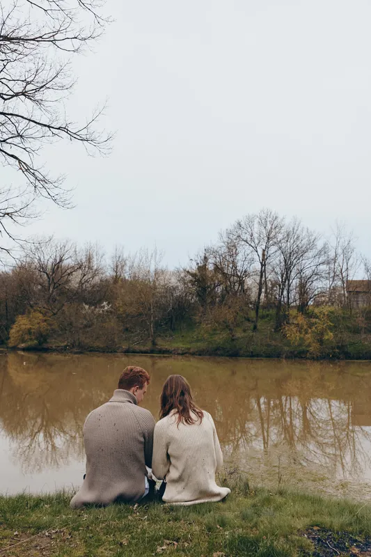 A young couple wearing cozy knitted sweaters sits together by a calm river on a chilly autumn day enjoying a peaceful moment in nature.