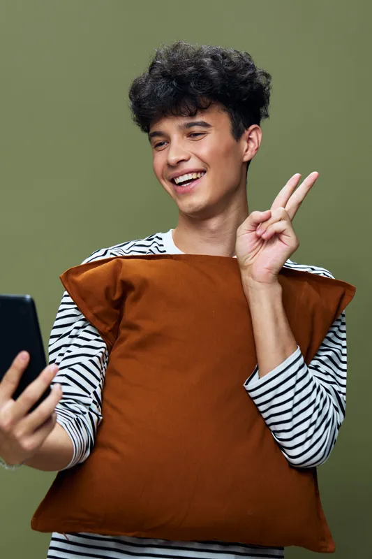 smiling young man holding a brown pillow and making a peace sign while taking a selfie against green background, casual striped shirt, cheerful mood