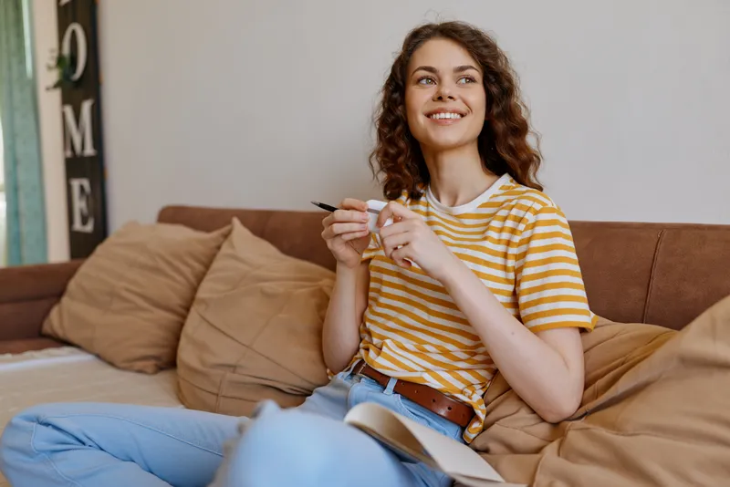 A smiling woman sits on a beige sofa at home, wearing a striped t shirt and jeans, holding a pen above a notebook. A bright, cozy living room frames a casual lifestyle scene.