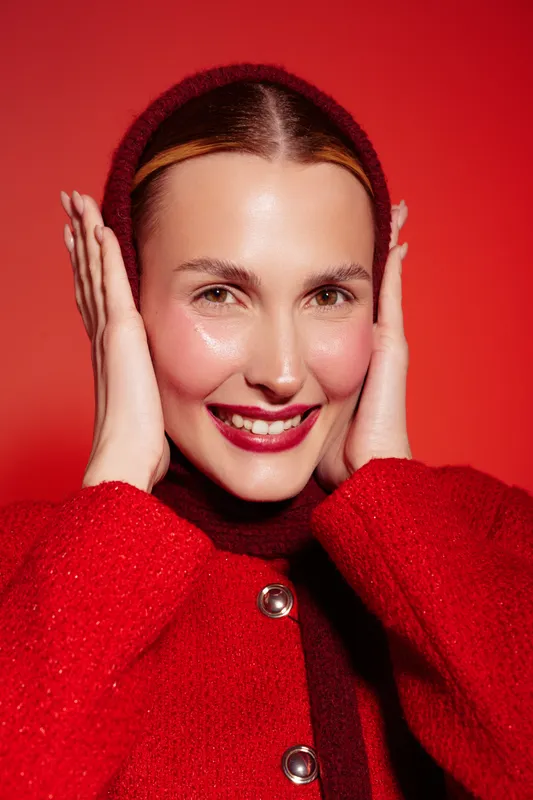 Smiling woman in red woolen coat and hat touches face with hands, posing on red background. Concept of winter fashion, warmth, and joyful mood in studio portrait.