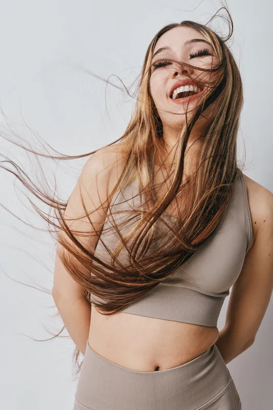 Young woman with long hair laughing and enjoying movement wearing beige crop top and pants, posing against plain background in casual fashion concept.