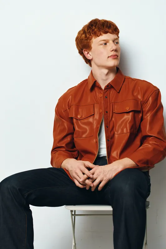 Young man with red hair wearing brown leather jacket sitting on white chair against white background looking aside with thoughtful expression in casual fashion style.