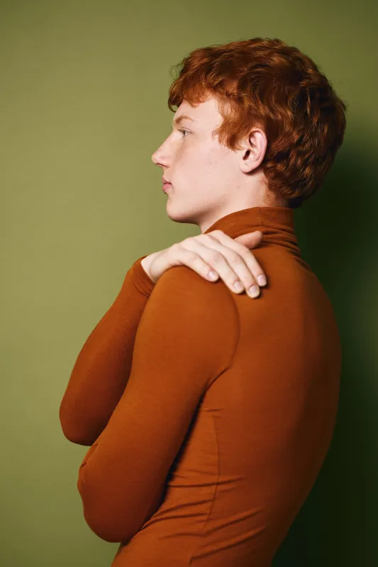 Profile of young man with red hair and freckles wearing brown turtleneck sweater looking ahead on green background. Side view, casual style and thoughtful pose indoors.