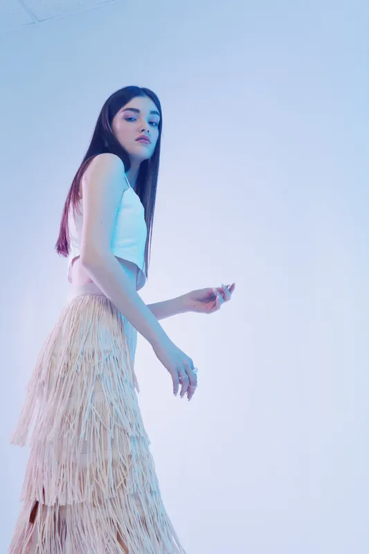 Young woman beauty fashion model in white top and layered fringe skirt posing with confident expression on light blue background. Studio shot with soft lighting for style concept.
