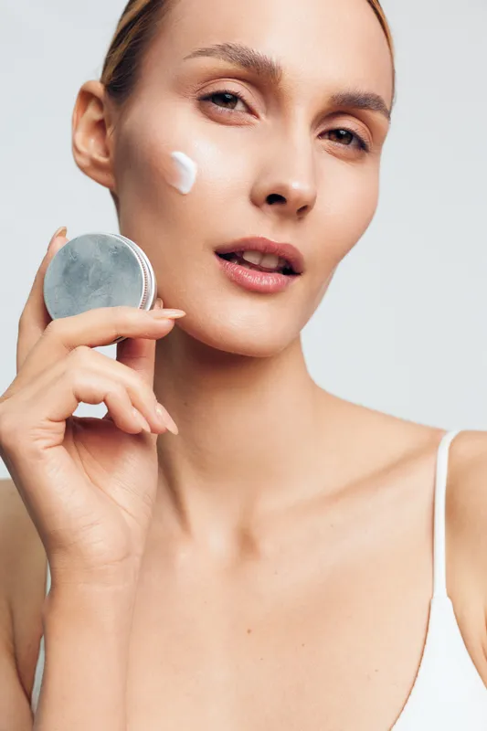 A young woman with clear skin applies white cream to her cheek using a round container. She looks directly at the camera with a neutral expression on a light background.