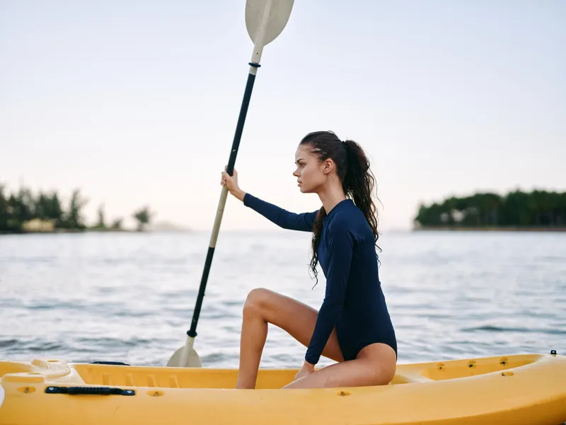 A lone woman sits in a yellow kayak on a calm lake, wearing a dark blue wetsuit, gripping a paddle upright as soft sunset light glows over the water.