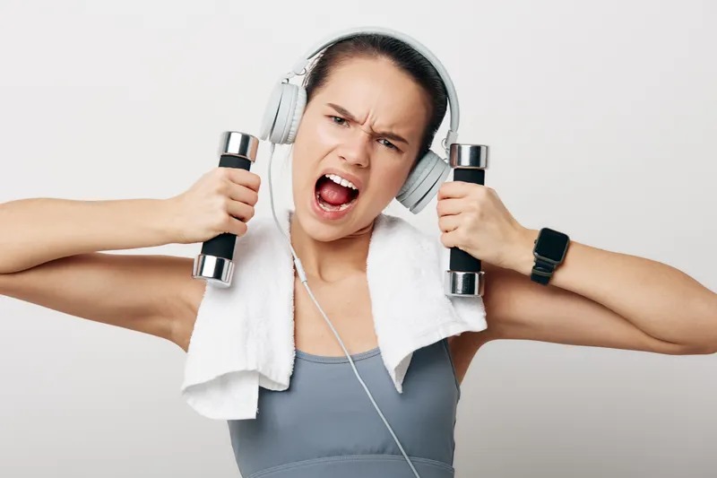 Fitness woman in workout gear lifts dumbbells with a towel over shoulders, wearing headphones, intense expression, ready for training, in a bright studio setting with clean backdrop.