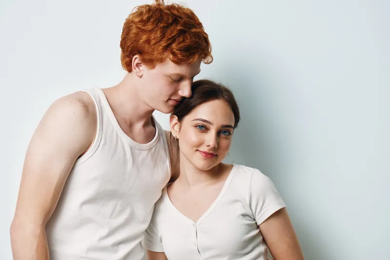 Couple, young man and woman, standing close together with affectionate gesture, casual white shirts, red hair, smiling and relaxed with white background for love and relationship.