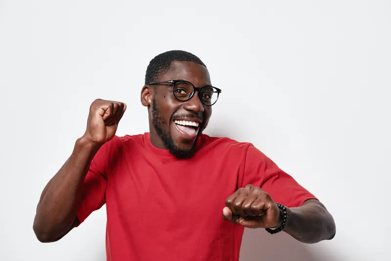 African American man with dark skin wearing glasses and red shirt smiling and dancing with raised fists on white background. Happy young black male in casual wear.