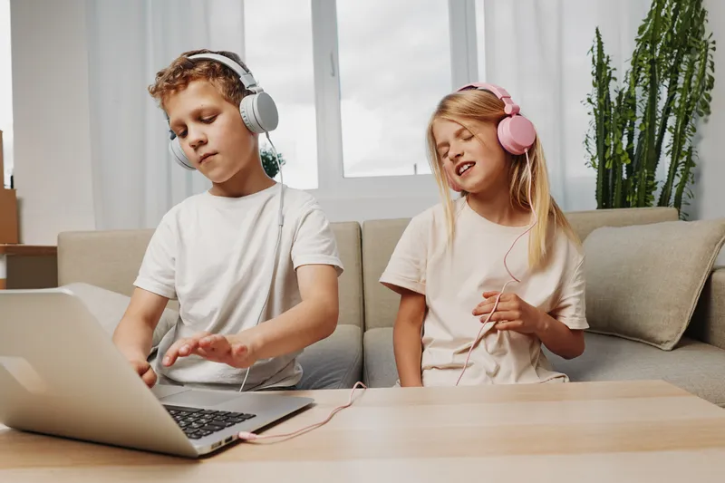 Boy and girl with headphones enjoy music together at home, using laptop on wooden table in bright living room with plants. Concept of childhood fun and technology.