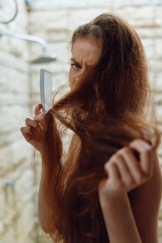 frustrated woman inspecting tangled dry hair while holding a white comb in a soft natural light bathroom setting, seeking hair care solutions