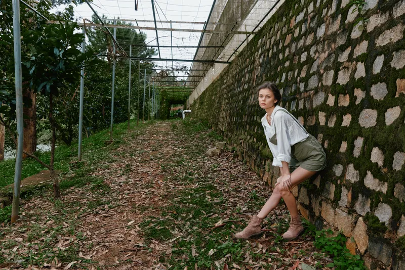 Young woman in a casual outfit sitting on a stone wall in a lush green environment, reflecting a carefree lifestyle and exploring nature s beauty