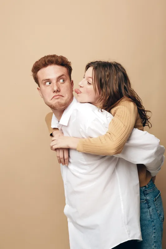 Young woman in beige sweater hugs surprised man in white shirt from behind on beige background. Concept of playful couple, affection, love, and relationship dynamics in casual style.