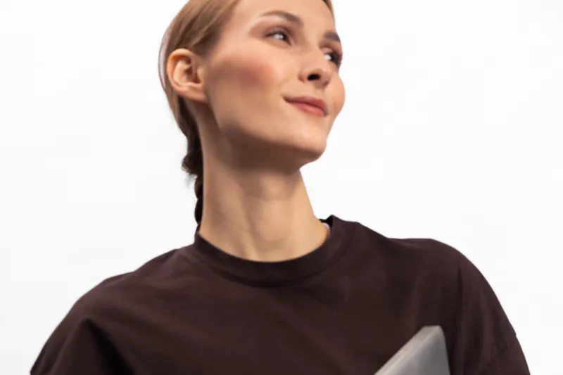 Confident young woman in dark brown shirt holds laptop and looks away with slight smile. Studio portrait on white background. Concept for technology, business, and modern lifestyle.