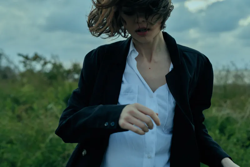Woman in a blazer and white shirt outdoors, wind in motion, fashion mood, natural light, field setting, candid style, atmospheric and stylish