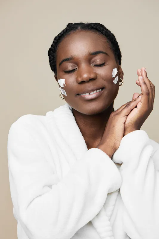 Young Black woman enjoying skincare routine with cream on her face, smiling and relaxing in a cozy white robe against a neutral beige background