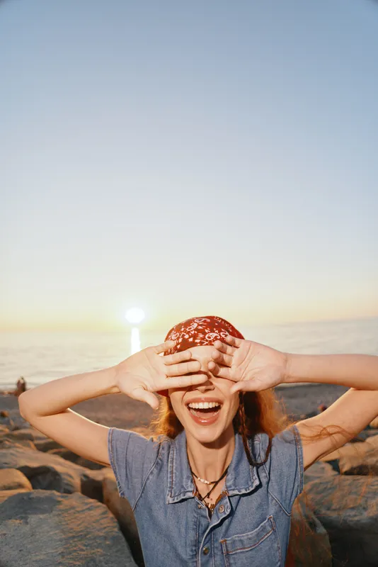 joyful woman in modern boho-western denim street style smiling and covering eyes on rocky beach at sunset with warm film color tones and casual accessories