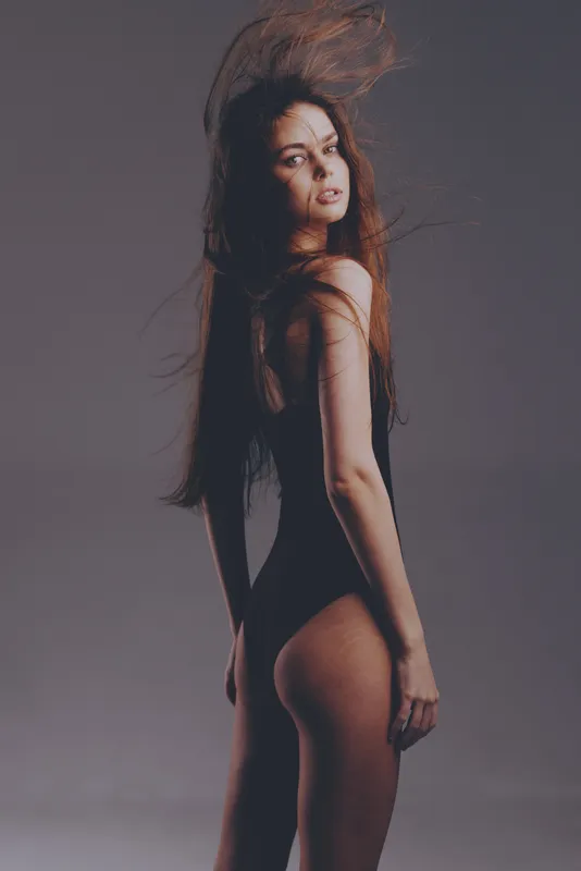 slim woman in black bodysuit posing with flowing hair on gray background, fashion, beauty, studio, elegant