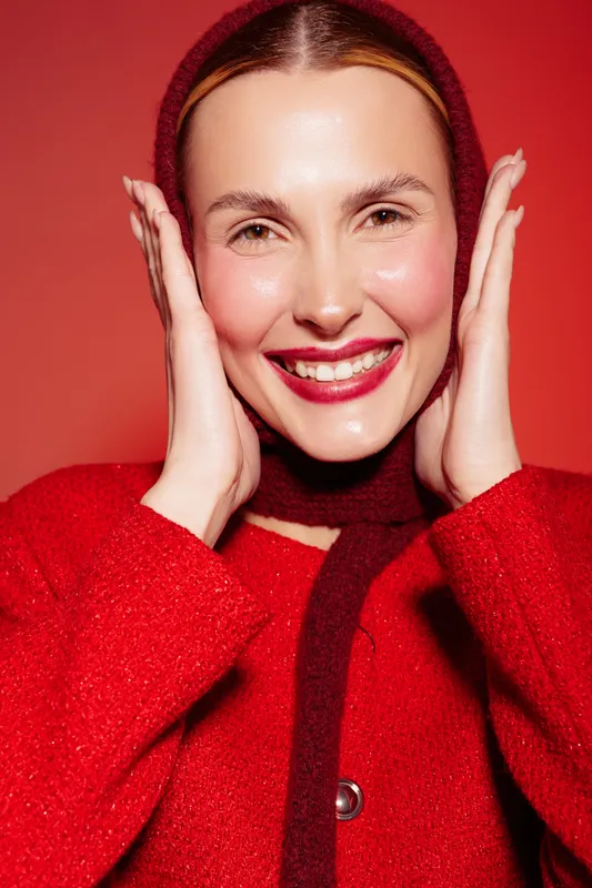 Smiling woman in red wool sweater and hood touches face with hands, showing natural skin glow and bright lipstick. Studio portrait on red background with warm lighting and happy mood.