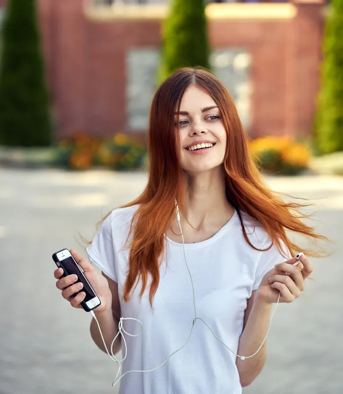 Женщина в белой футболке идёт по улице в наушниках с телефоном