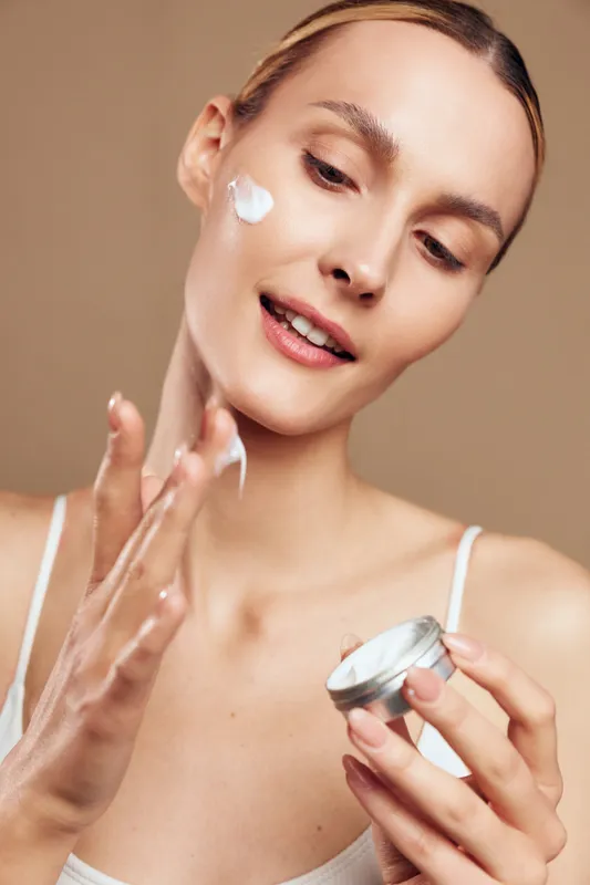 Young woman applies cream on face holding small jar with skincare product close to neutral background, demonstrating beauty routine and healthy skin care concept.