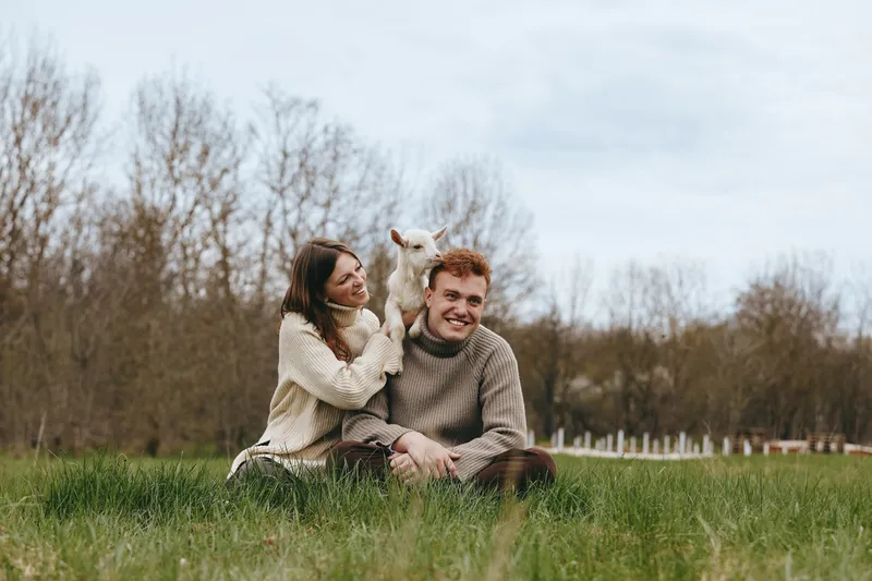 Smiling couple sitting on grass in a park with a playful goat kid perched nearby, sharing a warm moment in nature. Casual lifestyle scene highlights connection, happiness, and outdoor relaxation with