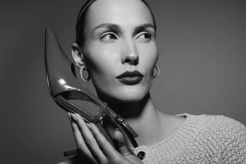 Young white woman with smooth skin holds pointed heeled shoe near face, looking aside. Studio portrait in black and white with elegant makeup and hoop earrings.