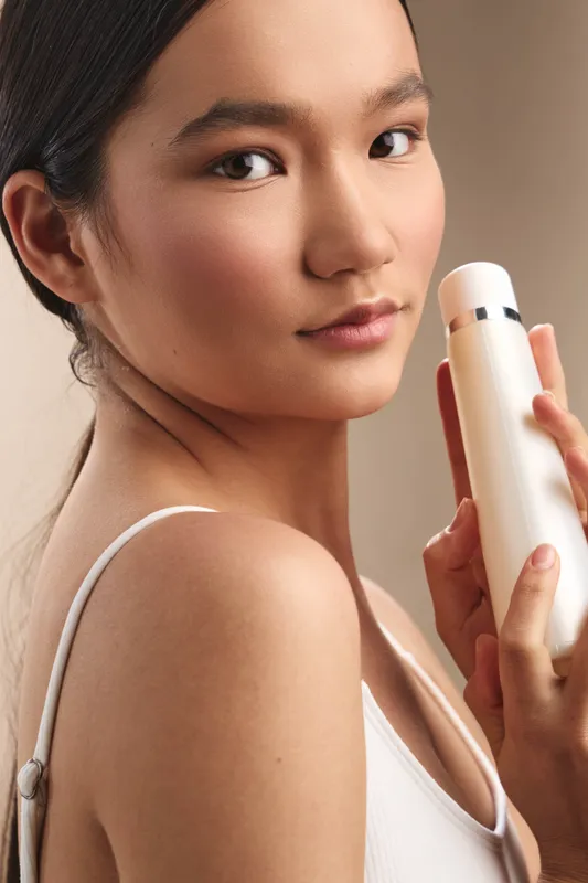 Asian woman models a skincare routine, glancing at the camera while holding a bottle. She wears a white top, in soft studio light that highlights healthy, radiant skin.