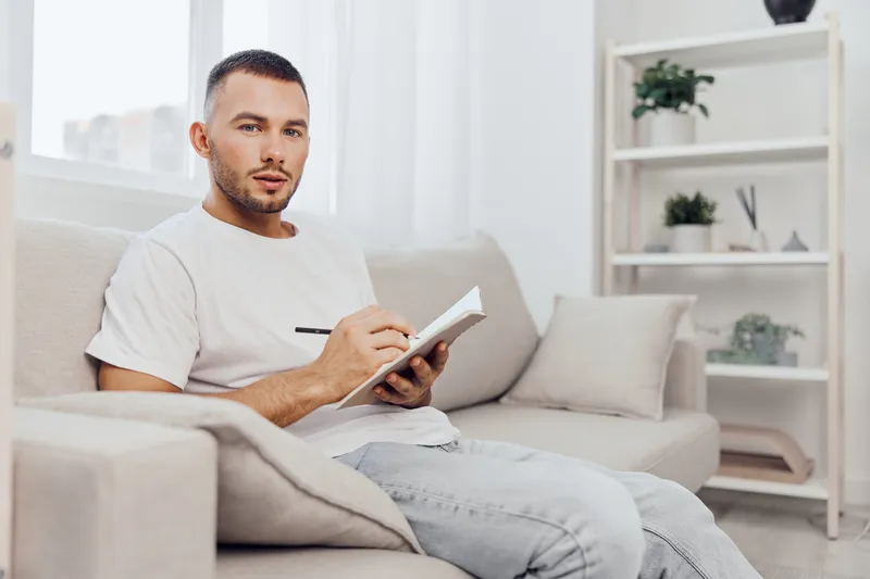 Thoughtful man sitting on a couch, writing in a notebook, surrounded by plants, capturing the essence of creativity and productivity in a cozy home environment.