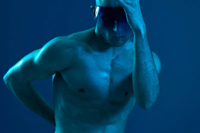 A thoughtful man with sunglasses poses against a blue background, highlighting his sculpted physique and emotional depth in a dramatic lighting setup.