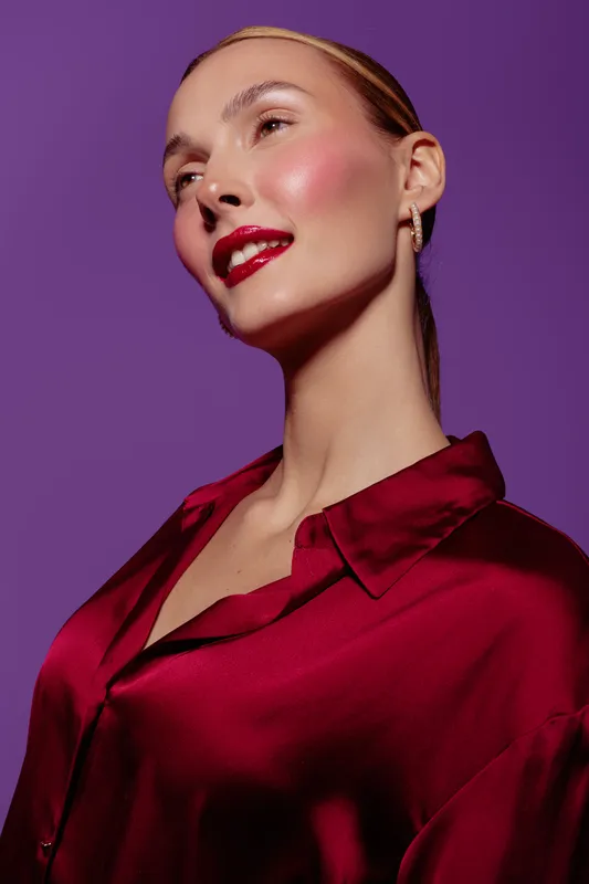 Portrait of confident woman with red lipstick and shiny skin wearing silky red blouse on purple background. Concept of beauty, fashion, elegance, and glamour.