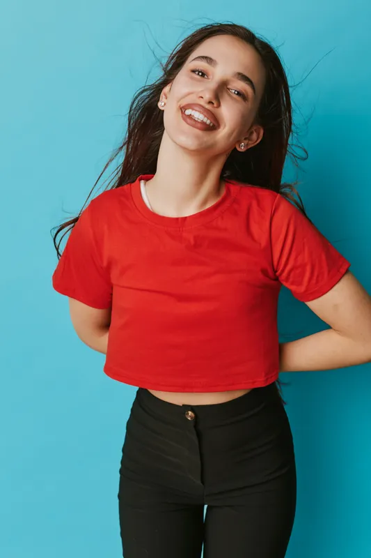 Young woman smiling with hands behind back wearing red crop top and black pants standing against blue background. Portrait of cheerful female with long dark hair and casual style.