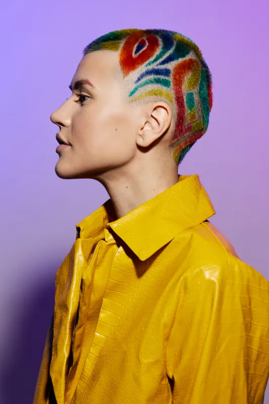 young woman with colorful shaved hairstyle wearing bright yellow jacket stands against vibrant purple background, showcasing bold fashion and creative hair design.