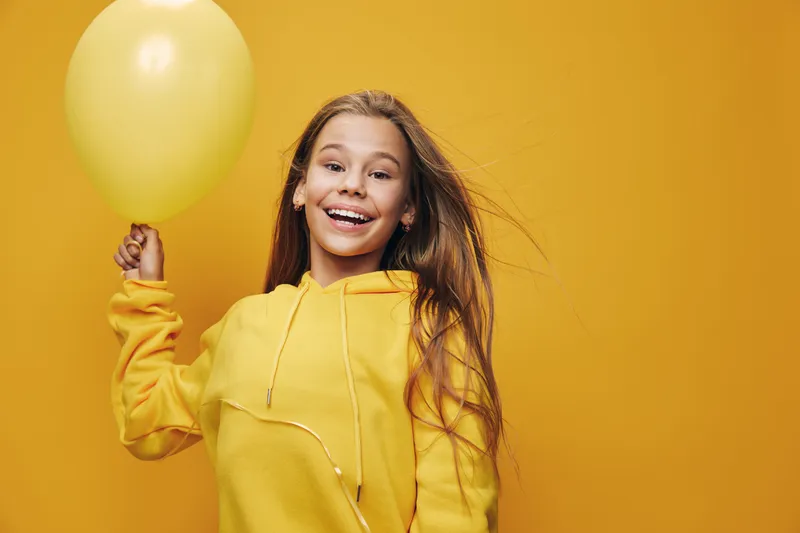 Girl smiling and holding yellow balloon wearing yellow hoodie on yellow background for celebration, happiness, joy and party mood with long hair and cheerful expression.