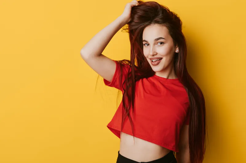 Young woman with long hair wearing red crop top posing with hand in hair smiling against yellow background. Casual style, beauty, confident expression and studio portrait.