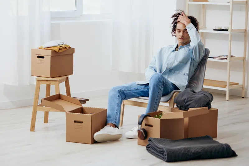 Парень с кудрявыми волосами распаковывает вещи из коробок в комнате, разбирая их