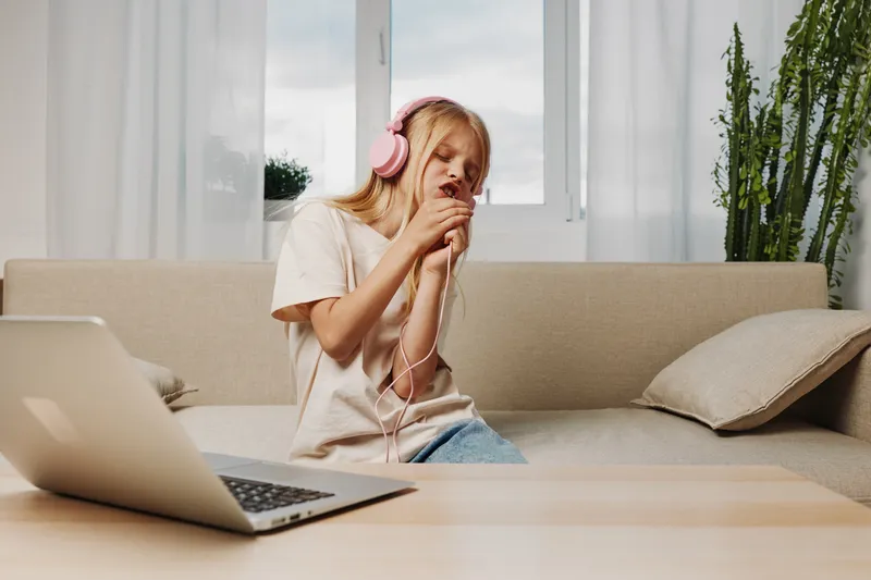 Young woman with pink headphones singing passionately while sitting on beige sofa in bright room. Laptop on table suggests casual home entertainment and music enjoyment concept.