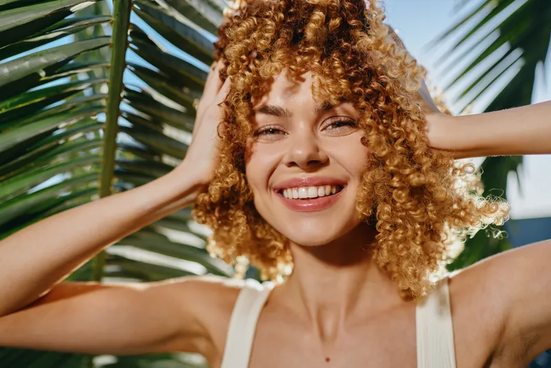 smiling woman with curly hair enjoying sunny tropical day near palm leaves, natural light, relaxed mood, healthy skin glow, summer vibe