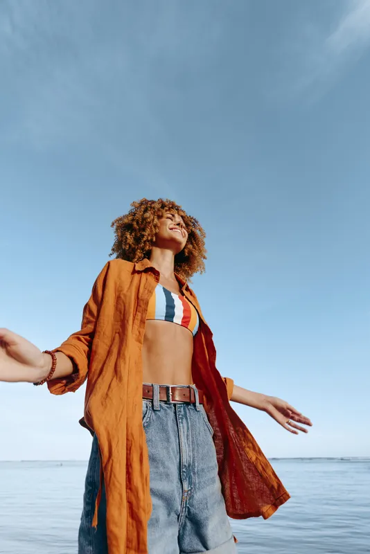 Young woman with curly hair standing by the sea, enjoying fresh air, blue sky background